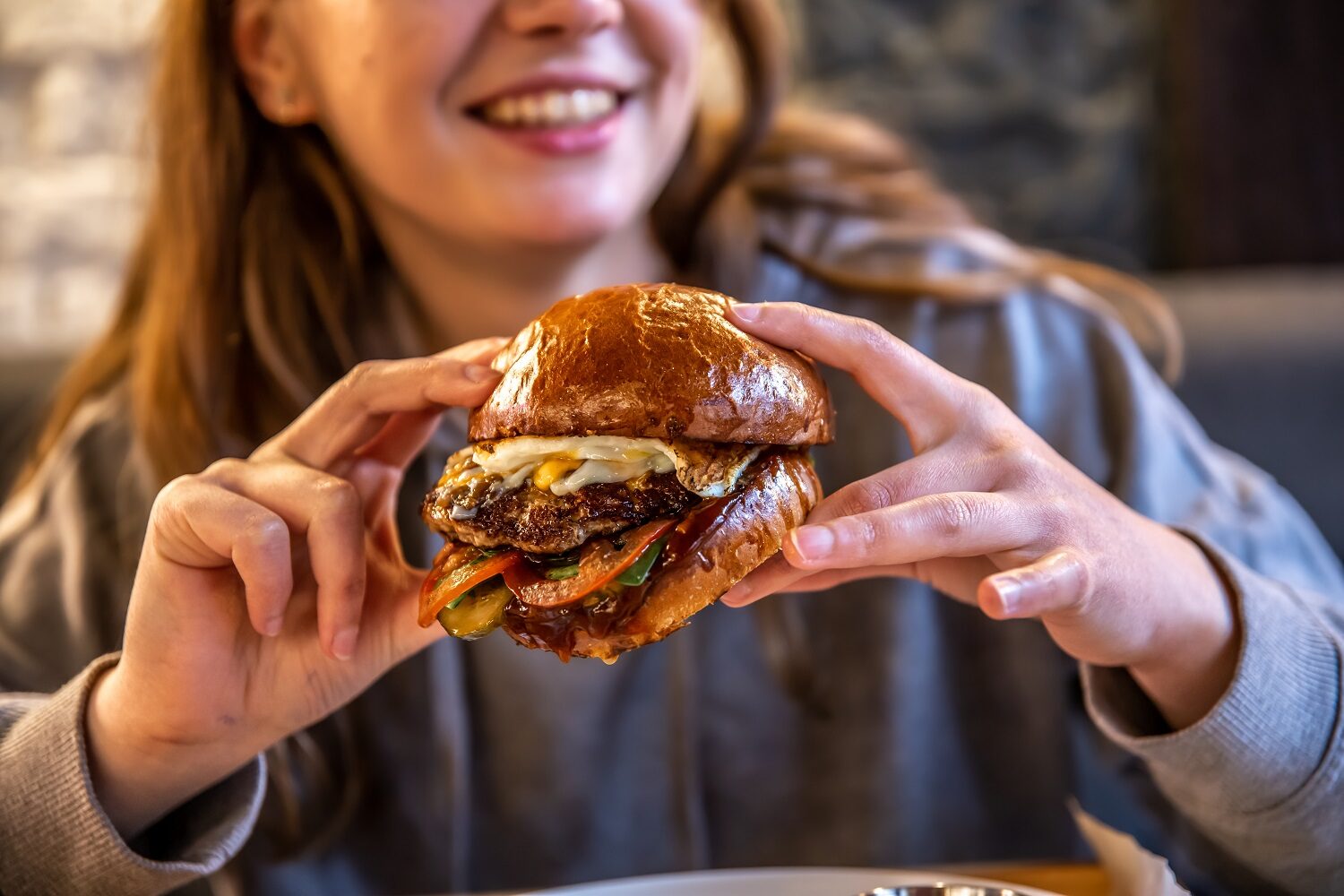 Close-up of a girl eating a big burger with meat and vegetables on a blurred background.