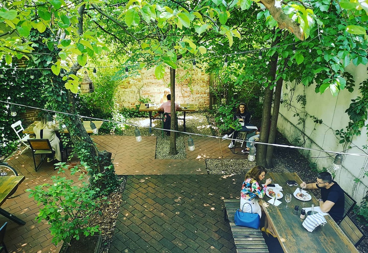 Grădina restaurantului Simbio din Bucuresti, privire de ansamblu, cu mese asezate printre copacii desi si mese care iau masa