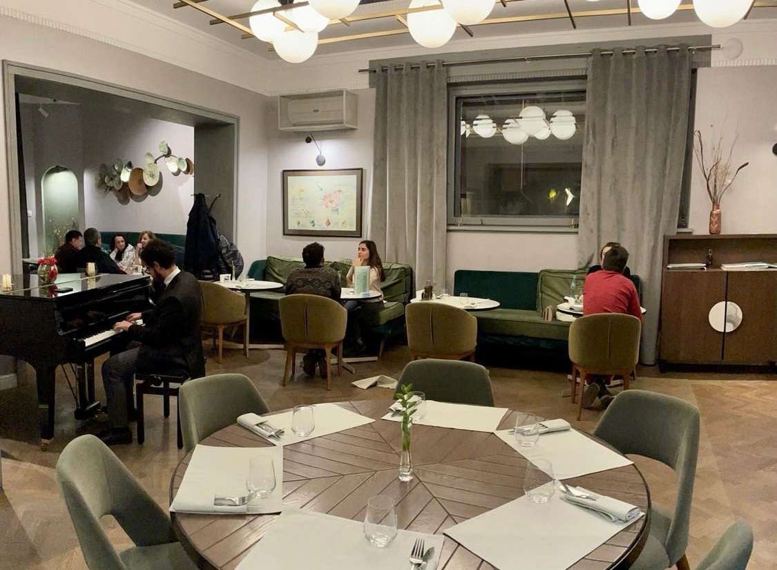 prim plan cu o masa rotunda, iar in fundal oameni asezati la mese si un barbat care canta la pian, la restaurant Savart București