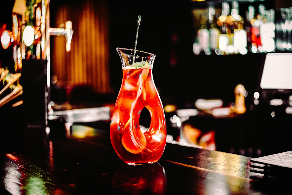 Carafa cu Sangria, de culoare rosi, prin care plutesc felii de citrice, pe fundalul inchis la culoare al unui bar.
