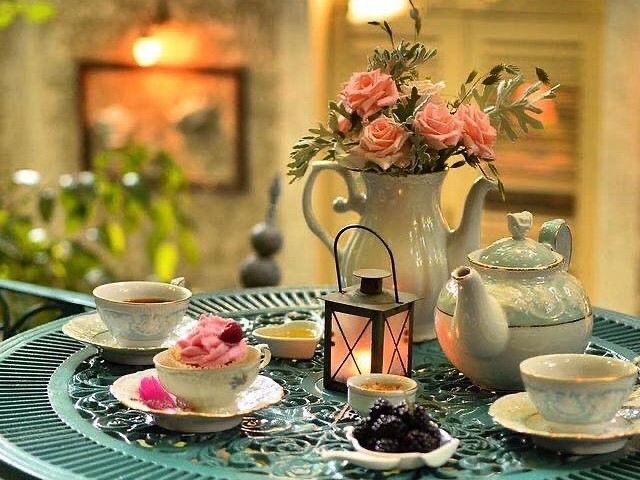 masa rotunda albastra din fier forjat, pe care sunt asezate 2 cesti cu ceai, o ceasca cu o prajitura, un felinar aprins, un ceainic si o vaza cu trandafiri roz, la ceainăria Ininitea București cana cu ceai de