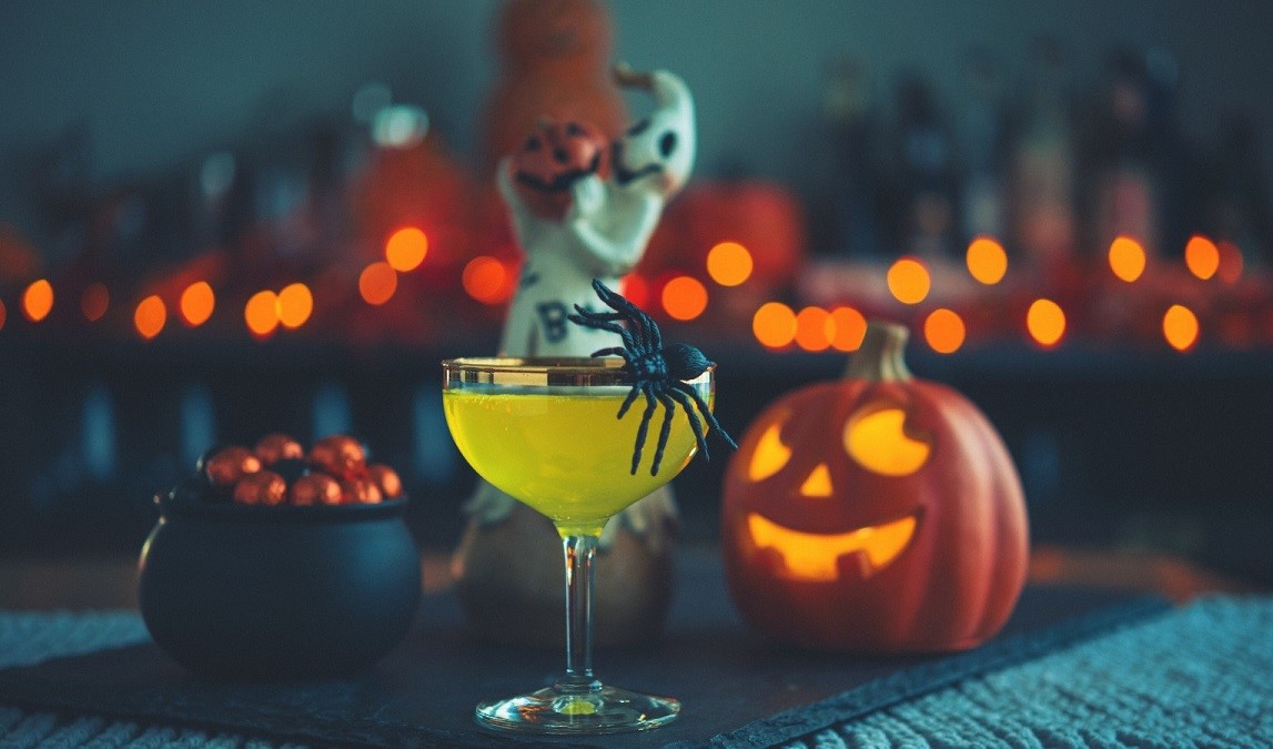 pahar de coktail cu un paianjen din fetru pe margine, iar in fundal un dovleac sculptat si luminat si un ceaun de vrajitoare plin cu bomboane, pe fundal inchis cu luminite, imagine pentru meniurile speciale de Halloween la restaurantele din București