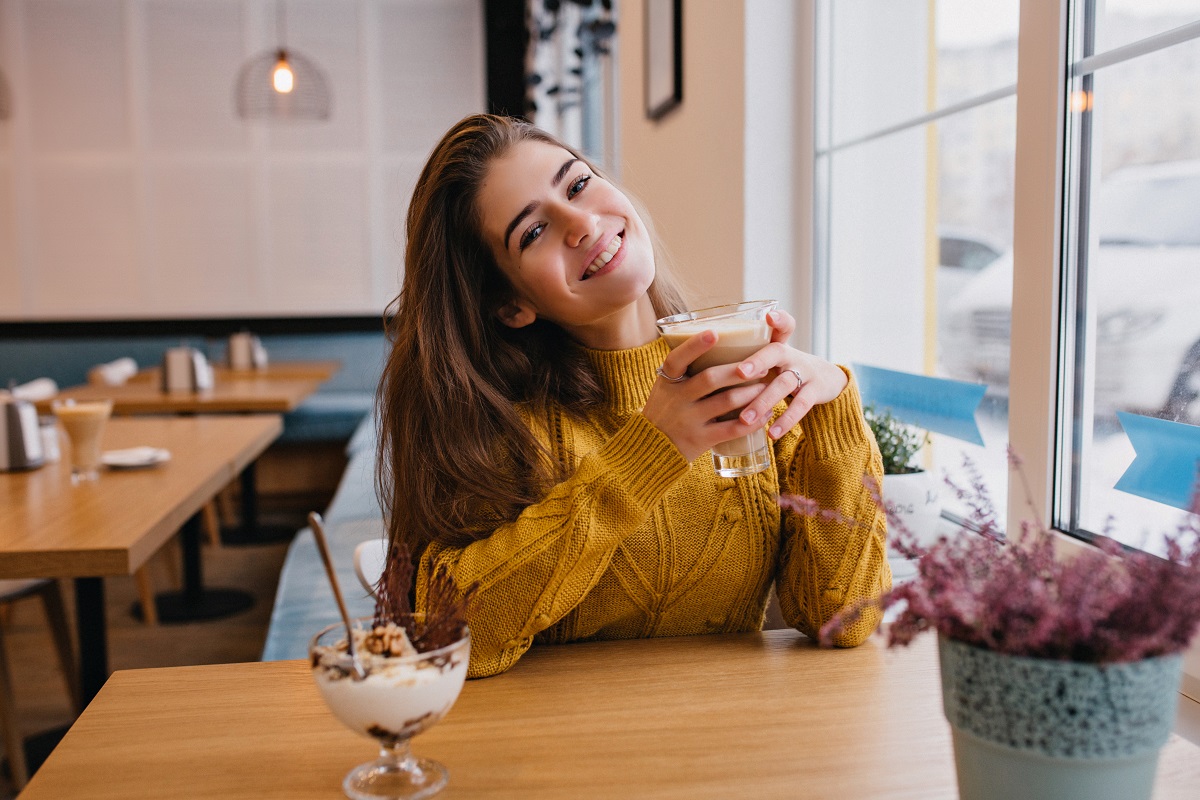 Pleased girl with dark hair chilling with cup of coffee in cozy cafe in winter. Indoor portrait of amazing lady in knitted yellow cardigan resting in restaurant and enjoying ice cream.