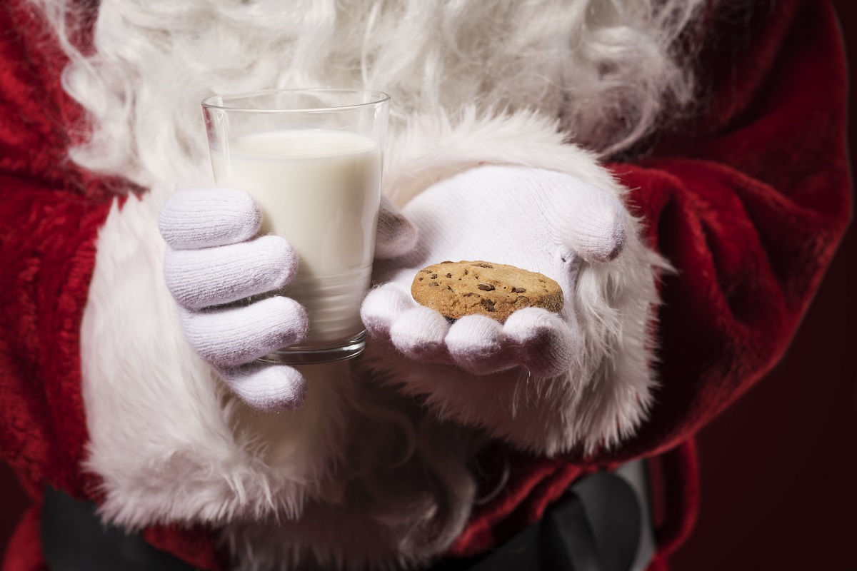 prim plan cu mainile lui Moș Crăciun cu manusi albe, in care tine un pahar de lapte si un biscuit