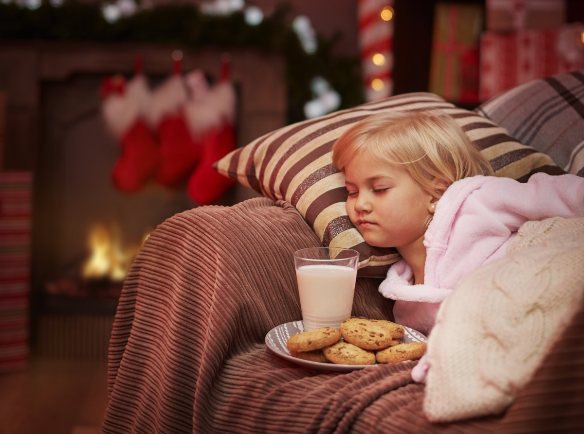 copil adormit pe canapea langa o farfurie cu un pahar de lapte și biscuiți pentru Moș Crăciun, iar in fundal ciorapi si luminite de Craciun
