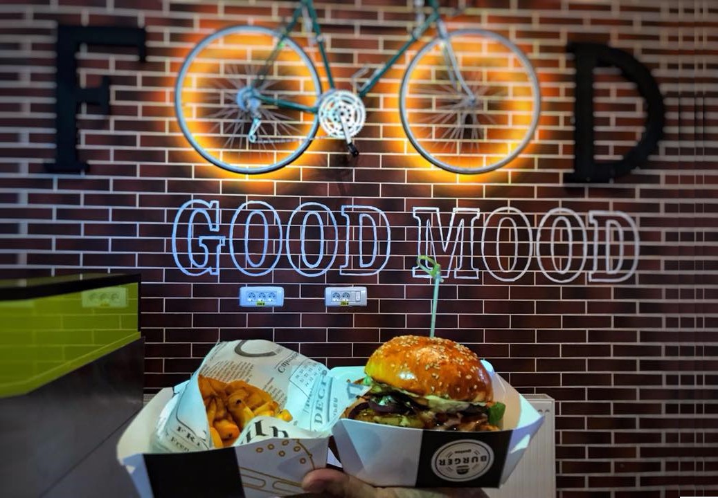 Un perete din caramaida pe care este atarnata o bicicleta cu roti iluminate si dedesubt e scris Good Mood, iar in prim plan o mana ce tine un burger si cartofi prajiti. Unul din cele mai bune restaurante din Sinaia 