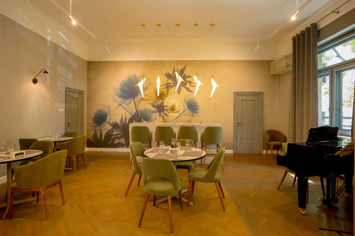 Incapere din restaurant Savart, unul din restaurante romantice București, cu un perete pictat cu flori mari, albastre, corpuri de iluminat moderne, o masa centrala, rotunda, cu scaune capitonate verzi, un pian si alte mese asezate pe laturile camerei