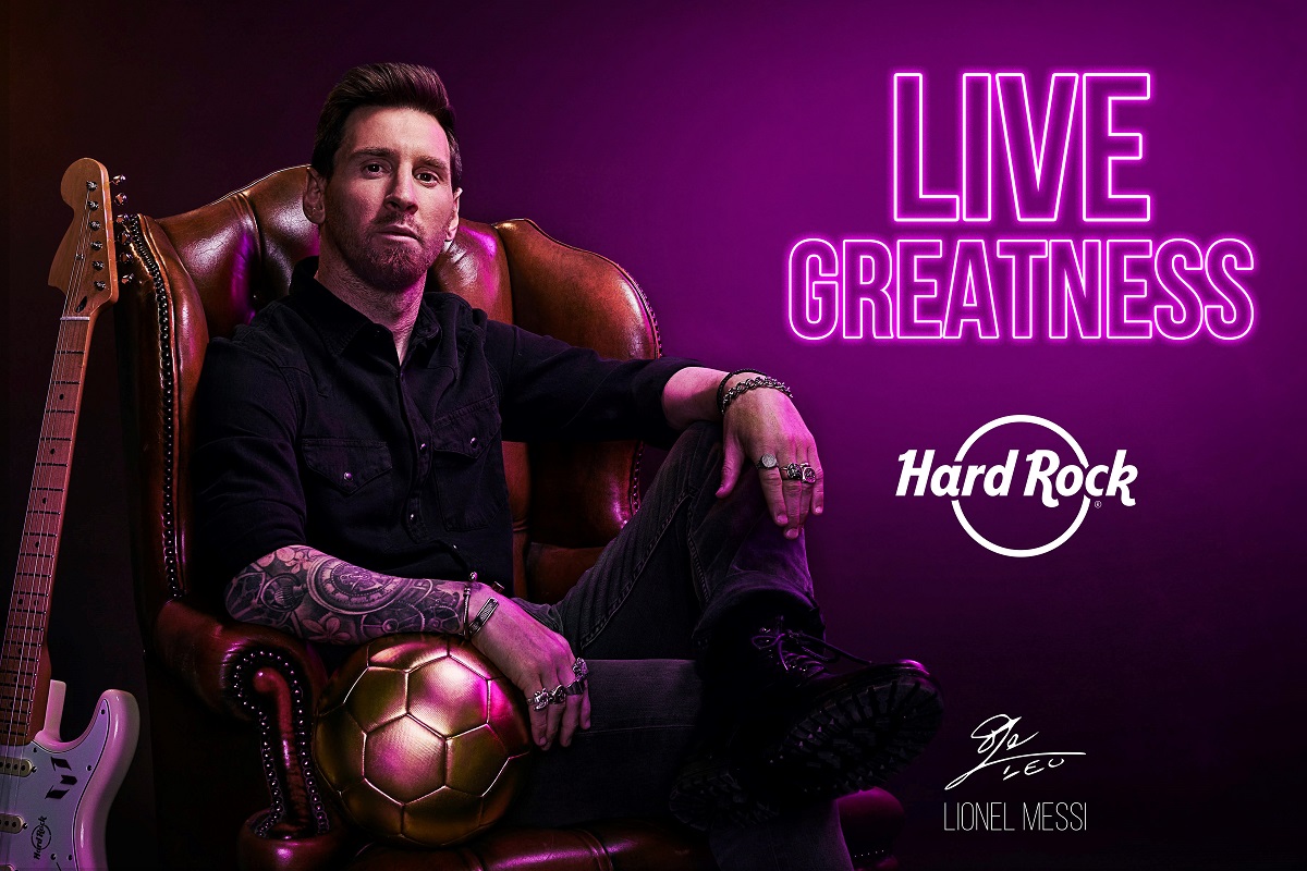 Lionel Messi fotografiat intr-un fotoliu mare de piele, pe fundal mov, pe care e sigla Hard Rock Cafe si lscris cu litere luminoase Live Greatness