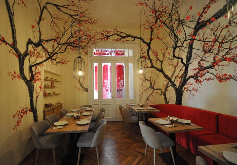 Sala de mese de la restaurant chinezesc PengYou Hala Traian, cu peretii decorati cu copaci de cires pictati, o fereastra mare, mese si canapele rosii