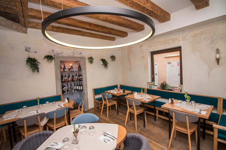Încăpere din Restaurant Dei Frati din Brașov, cu mese mici, pereti albi, un corp de iluminat mare, rotund, si tavan cu barne din lemn