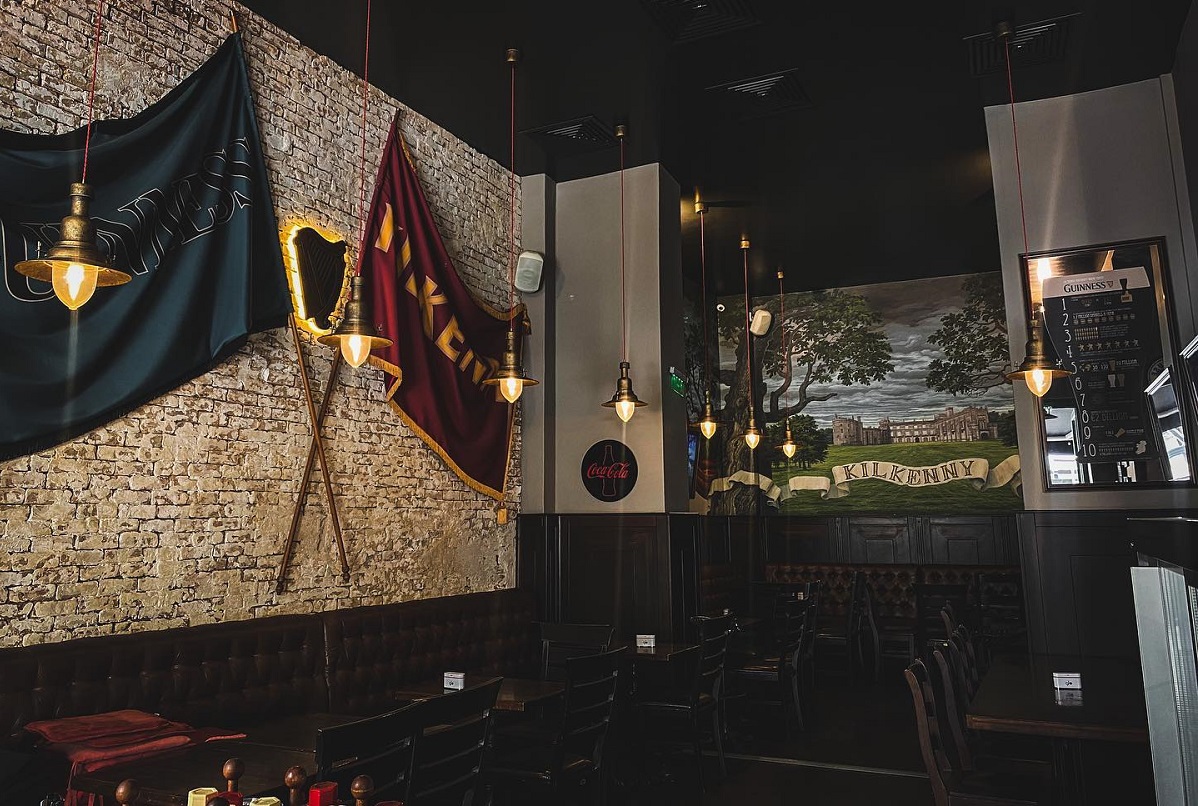 Kilkenny Irish Pub, cu mese si scaune din lemn maro inchis, pereti cu caramida aparenta alba, doua steaguri atarnate pe pereti, rosu si albastru, iar in fundal o pictura murala cu o pajiste si castelul Kilkenny