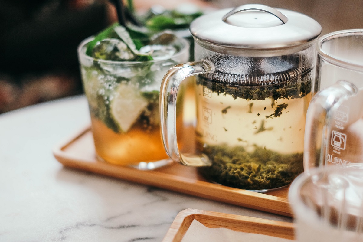 closeup cu o carafa in care se infuzeaza ceai si o cana de ceai transparenta langa ea cu ceai verde, ca parte din dieta antistres