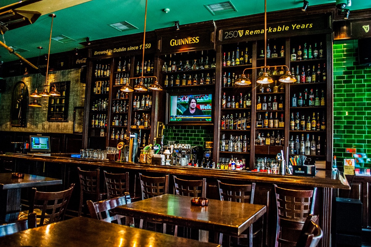 St. Patrick Irish Pub din București: imagine cu barul plin cu rafturi de bautura pana sus, masa de bar cu scaune inalte si in prim plan mese mici de lemn masiv