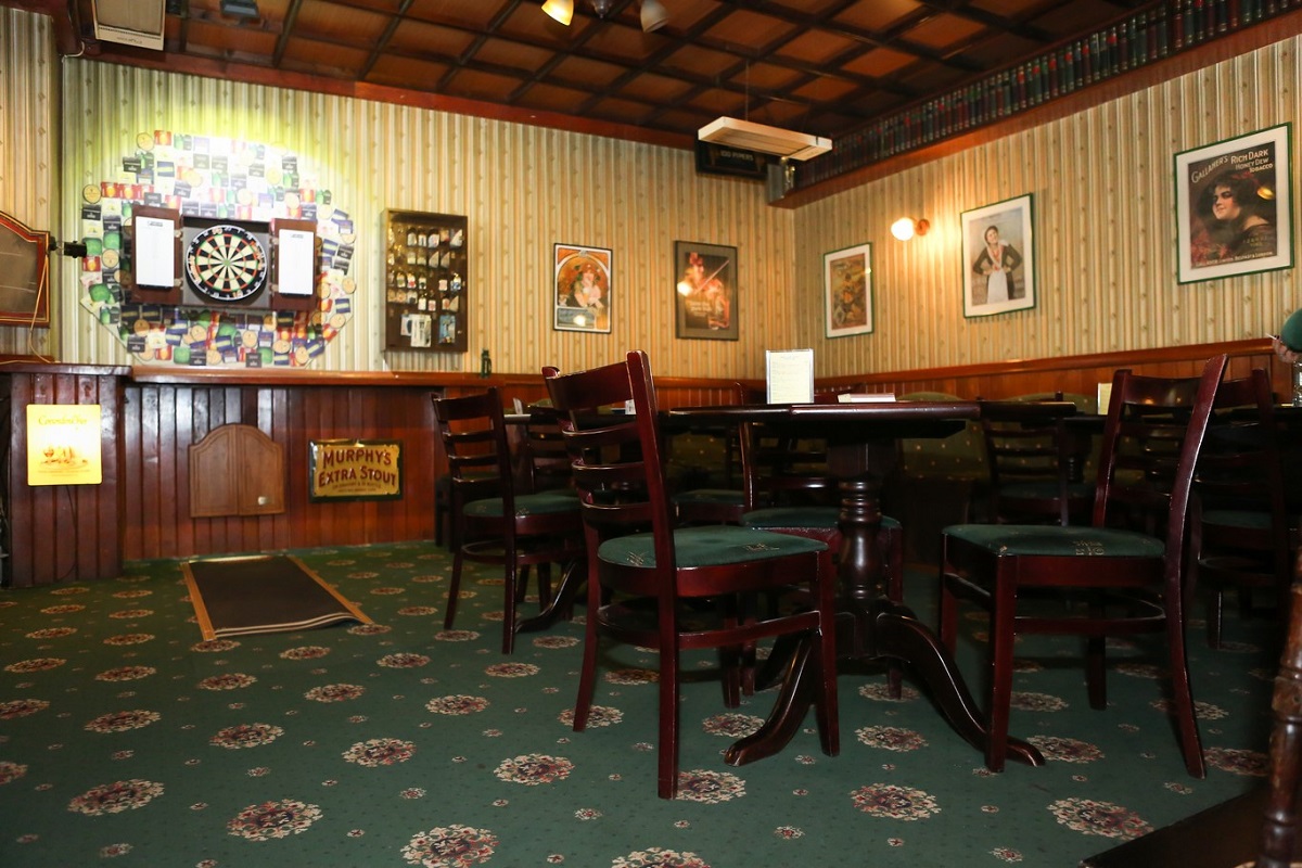 Salon din The Dubliner, unul din pub-urile irlandeze din București, cu covor verde inchis cu motive floarele, mese csi scaune din lemn maro inchis, pereti cu lambriu din lemn, tablouri si un panou de darts