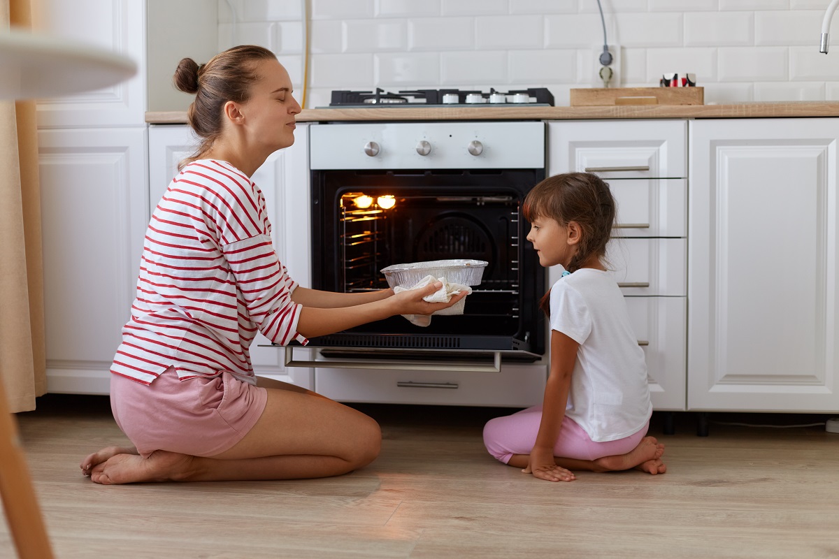Portrait of happy young woman taking baking out of oven, her daughter looking at tasty sweets, people wearing casual clothing, sitting on floor in kitchen, cooking together. Imagine reprezentativa pentru gustul copilăriei