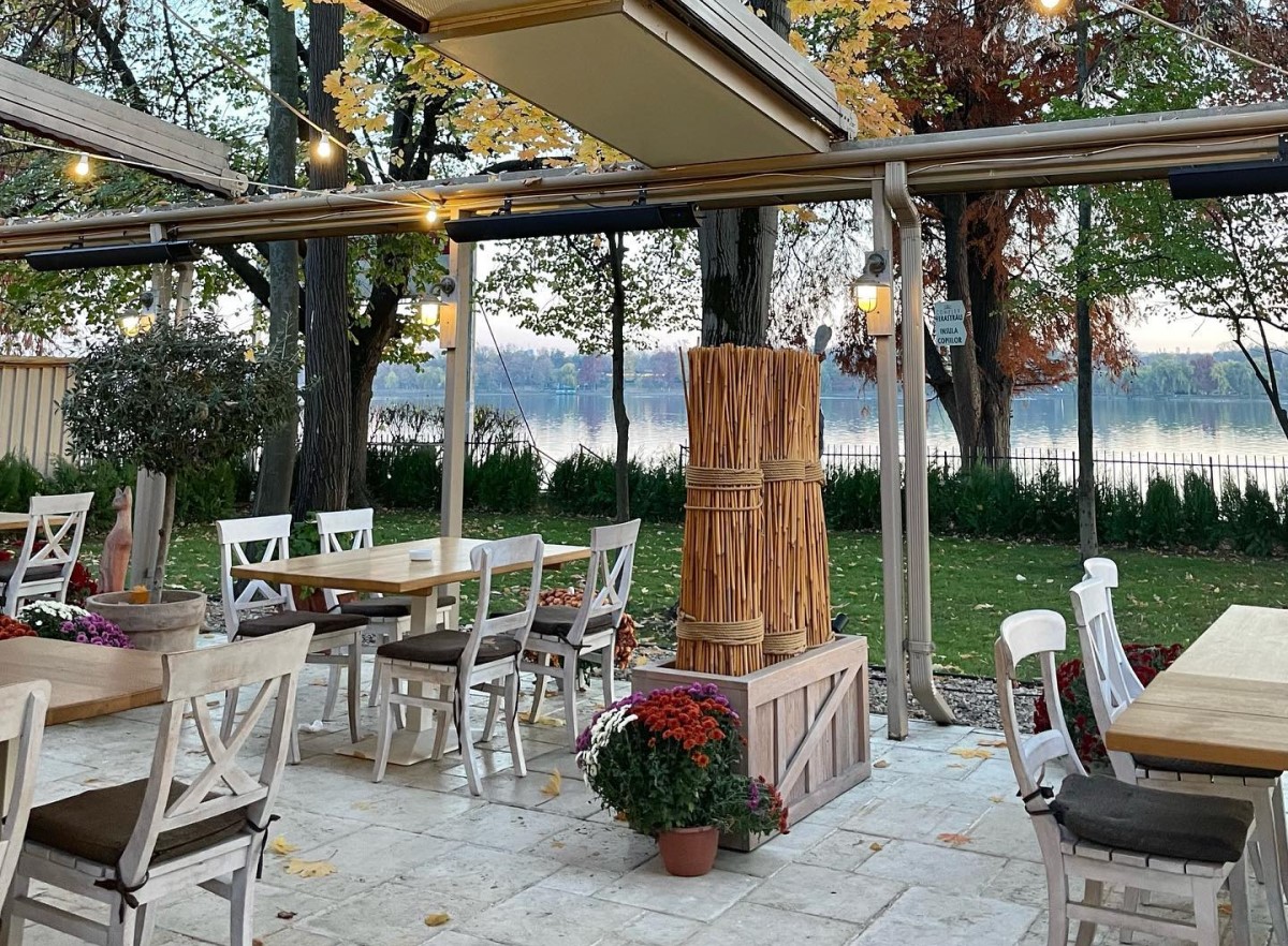 terasa de la restaurant Ancora din Bucuresti, cu mese din lemn si scaune albe, un manunchi de stuff ca decor, gazon si lacul Herastrau ce se vede in fundal