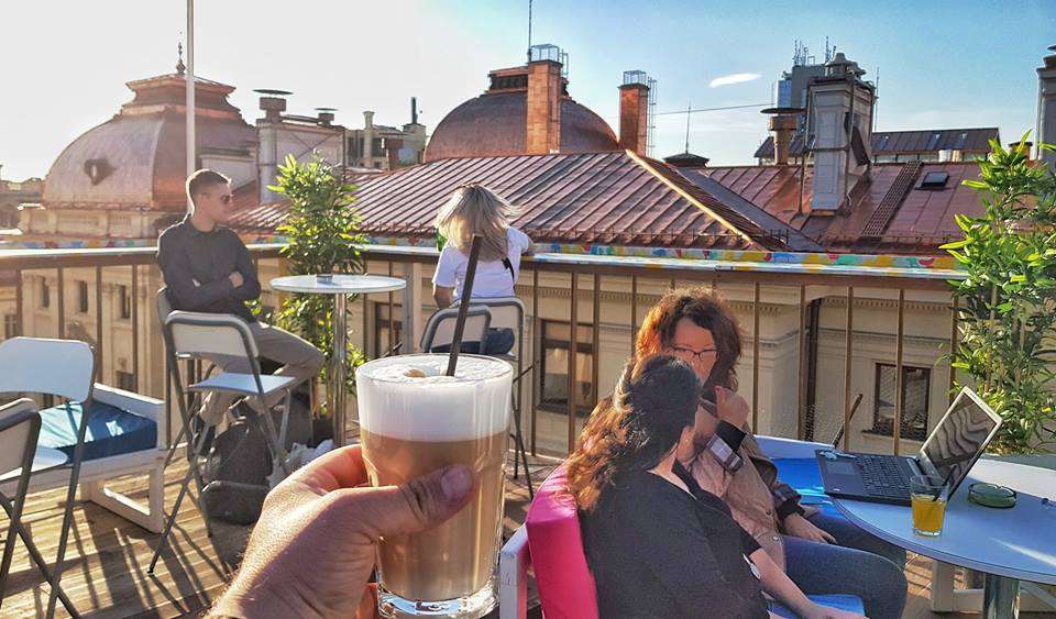 prim plan cu un pahar de frape tinut in mana, iar in fundal oameni care stau le mese in soare si acoperisurile caselor vecine de la Pura Vida Sky Bar București