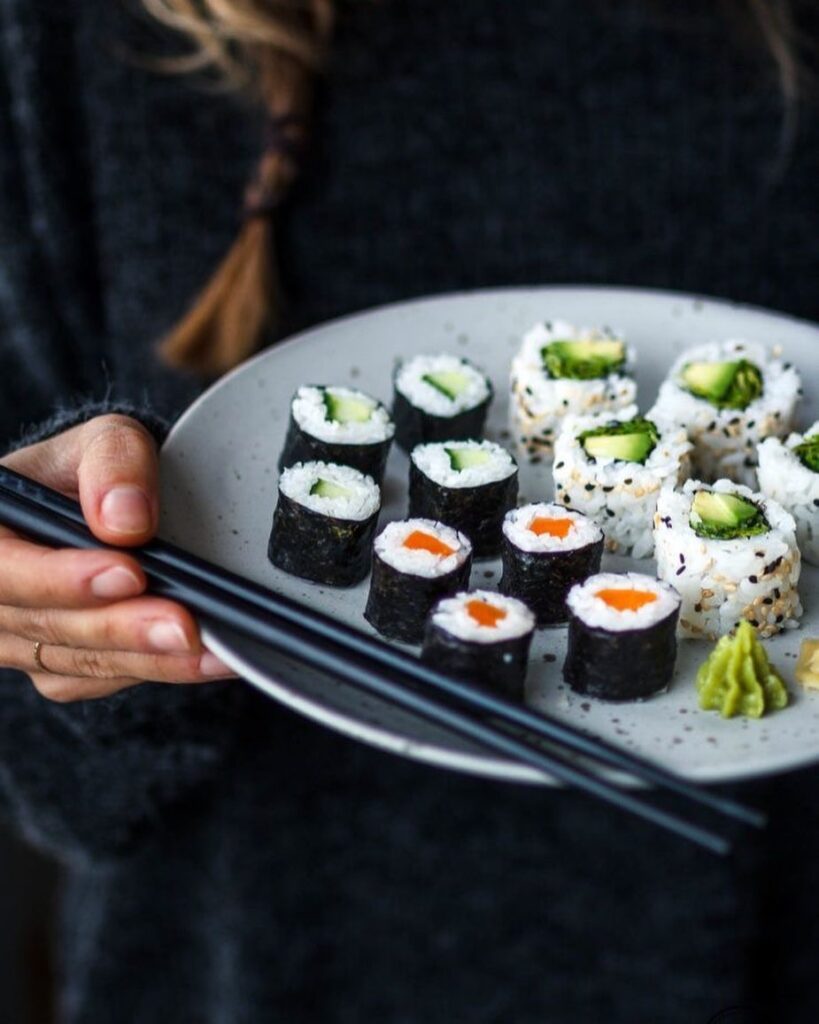 closeup cu o farfurie cu sushi tinuta in maini de cineva, alaturi de betisoare japoneze