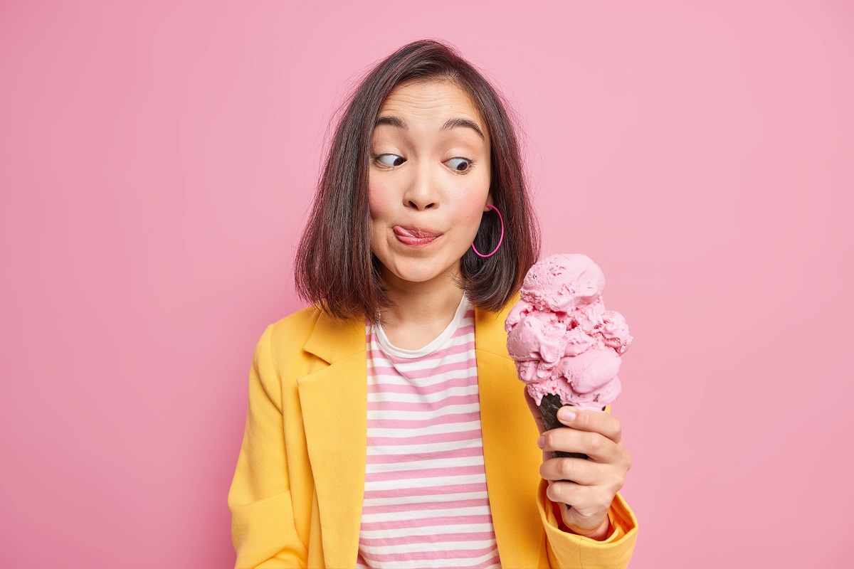 Pretty young dark haired Asian woman looks at appetizing yummy ice cream licks lips with tongue cannot wait until eating delicious summer dessert dressed in fashionable clothes. Sweet tooth concept