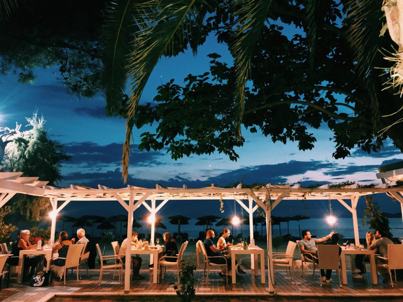 Restaurant San Atonio din Potos, Thassos, fotografiat seara, cu o pergola din lemn si marea in fundal, mese asezate dedesubt si oaspeti care iau masa