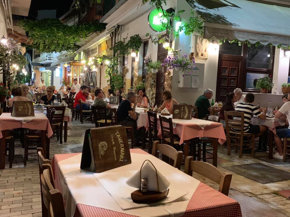 Taverna Tavernaki din Thassos, fotografiata seara, cu mese asezate pe o straduta ingusta cu flori si oameni asezati la mese