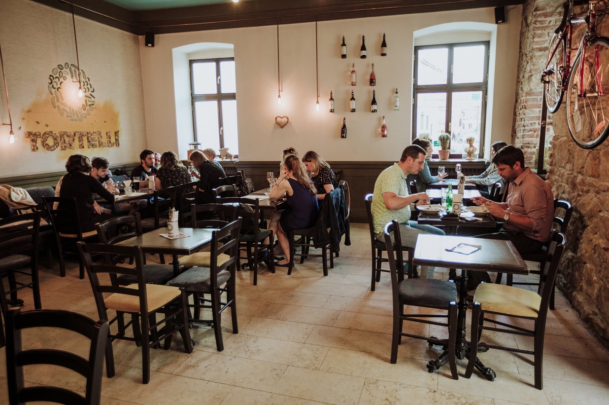 imagine de ansamblu din restaurant Tortelli Pasta Bar unde mănânci bine paste in Cluj, amenajat cu mese din lemn, cu oameni care iau masa, si ferestre in fundal