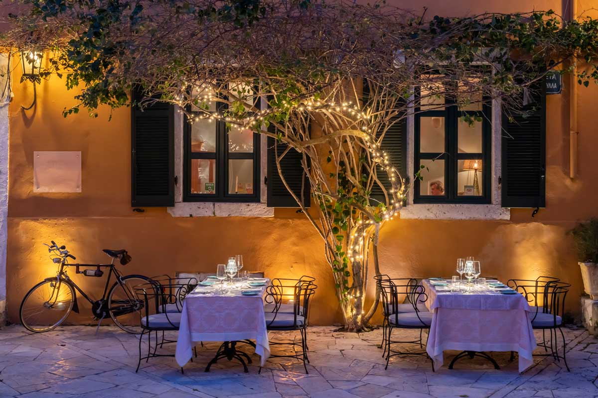 doua mese elegante cu lumanari pe masa asezate in dreptul unei case cu ferestre verzi si obloane si umbrite de un copac la Venetian Well restaurant de fine dining din Corfu