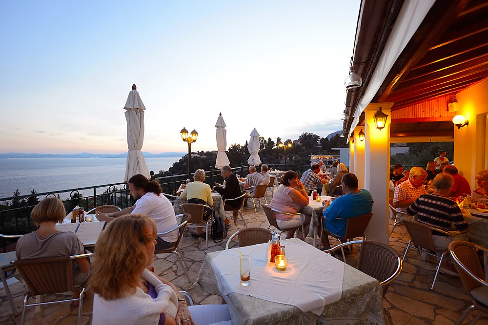 terasa Vitamins Taverna din Corfu, amplasata la inaltime, pe maulul marii, cu oameni asezati la mese, felinare pe pereti, podea pietruita si umbrele stranse