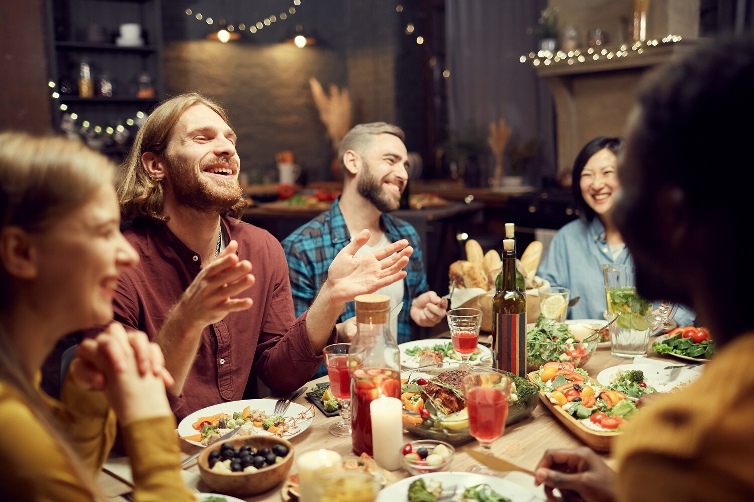 grup de tineri adunati in jurul mesei, razand fericiti, intr-o camera cu lumina redusa, imagine reprezentativa pentru mâncare pentru o seară cu prietenii