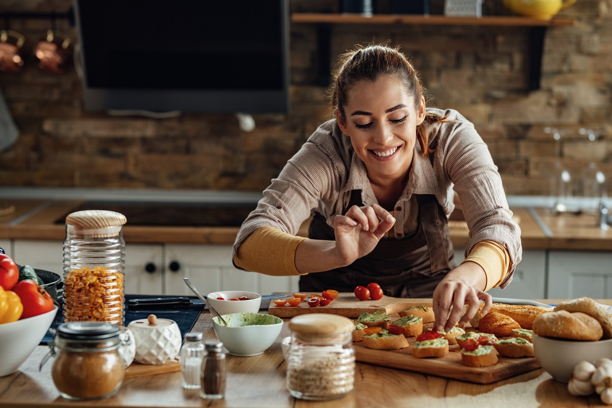Young smiling woman making bruschetta with healthy ingredients while preparing food in the kitchen.