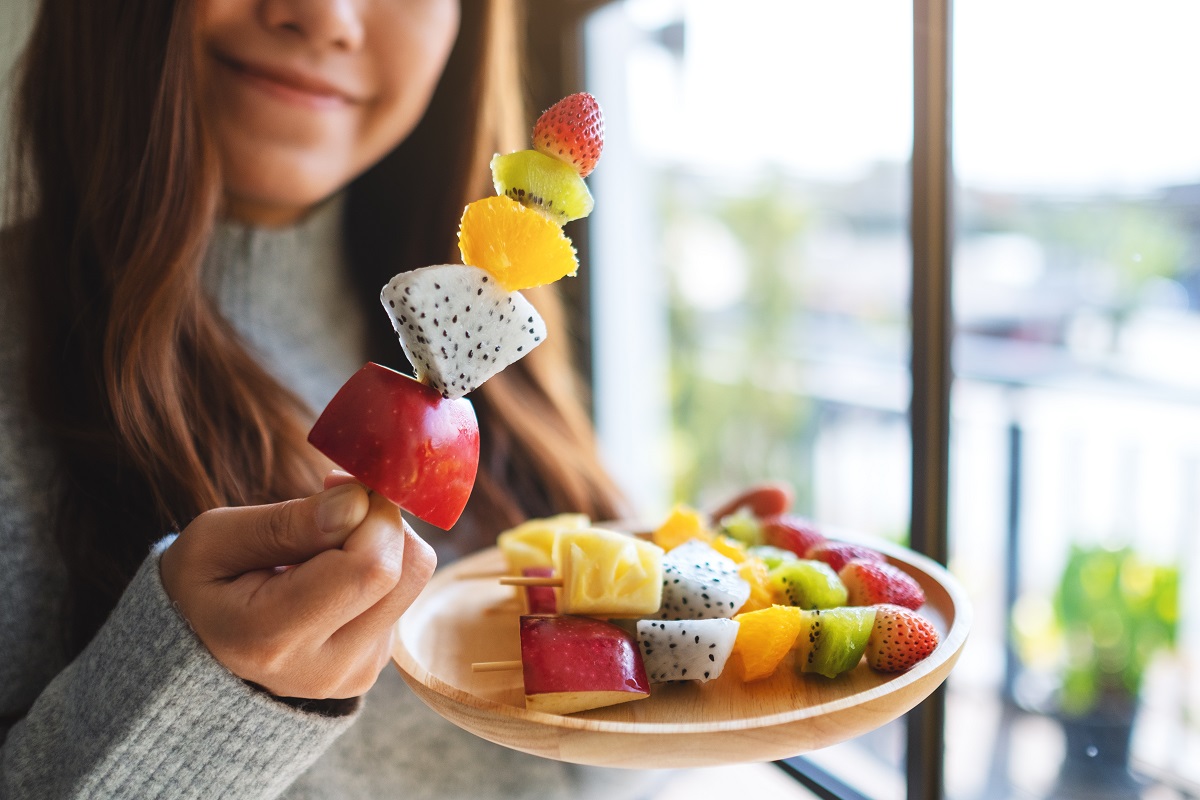 Closeup image of an asian woman holding and eating a fresh mixed fruits on skewers. 