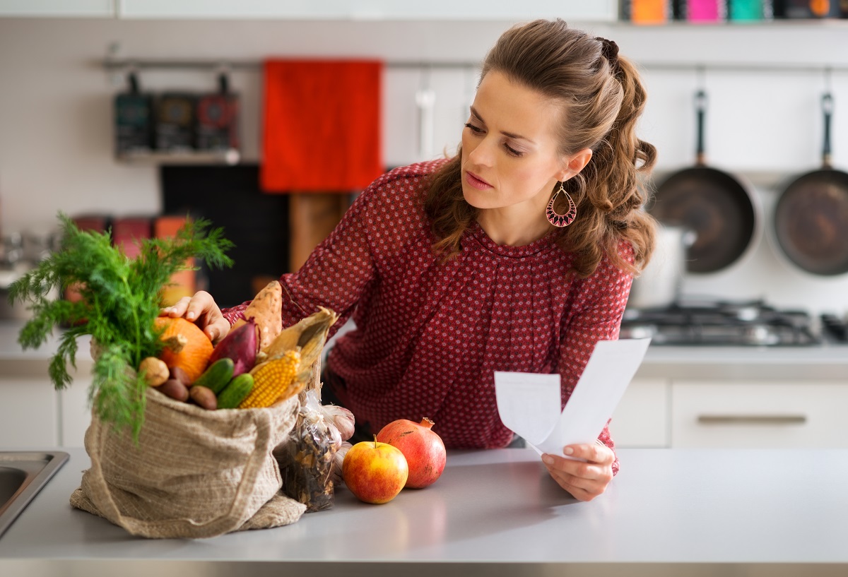 An elegant woman is leaning on the kitchen counter, checking the contents of her burlap bag which is filled with fresh autumn fruits and vegetables. She is double-checking that she has everything.