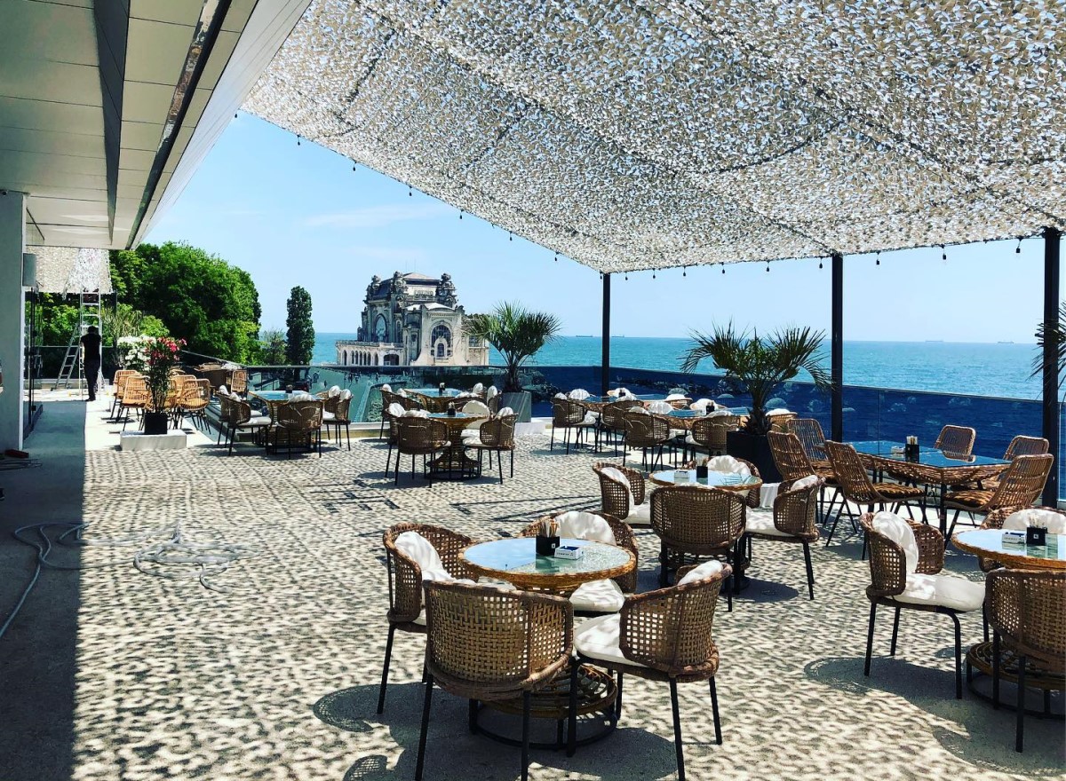 terasa pe malul marii, cu copertina alba si mese cu scaune din aratn, iar in fundal Cazinoul din Constanta, la restaurant The View