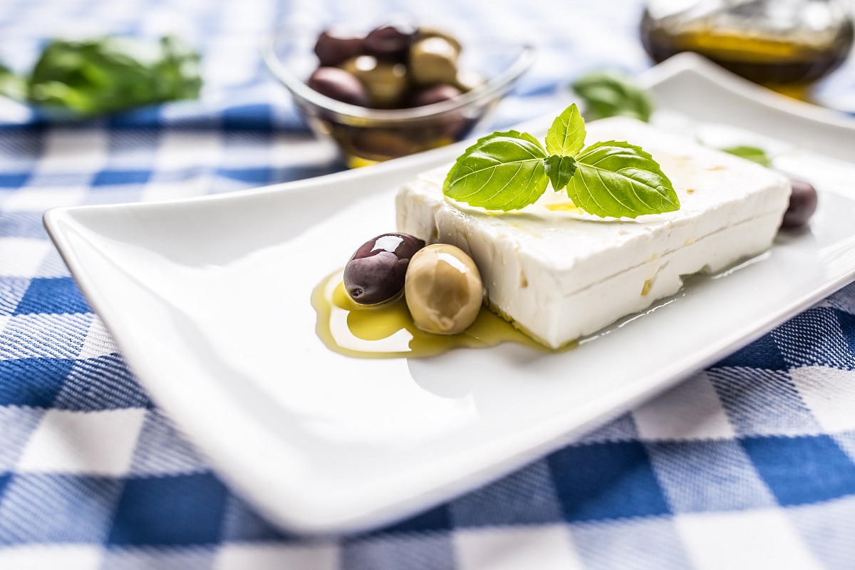 platou alb cu brânză grecească feta, cu ulei de măsline, alături de măsline, decorată cu o frunză de busuioc, pe o fata de masa in carouri alb albastre