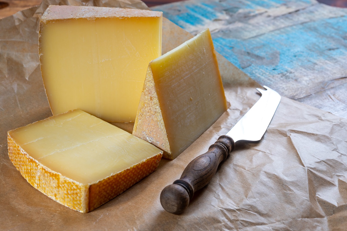 brânză Gruyere, elvetiana, asezata in bucati mari, triunghiulare, pe un platou de lemn alaturi de un cutit de brâzneturi