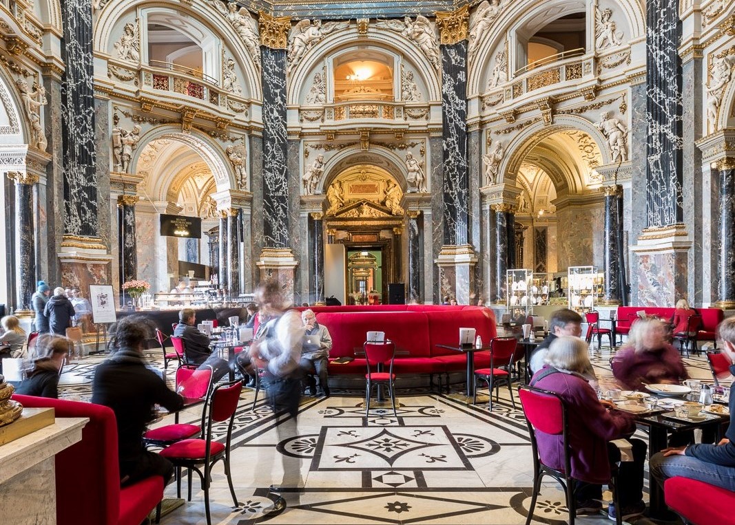 Kunsthistorisches Museum Cafe, o cafenea situata in interiorul muzeului de arta din Viena, cu interior somptuos, canapele din catifea rosie si oameni care iau masa. Cina la Muzeu, una din experiențe culinare inedite