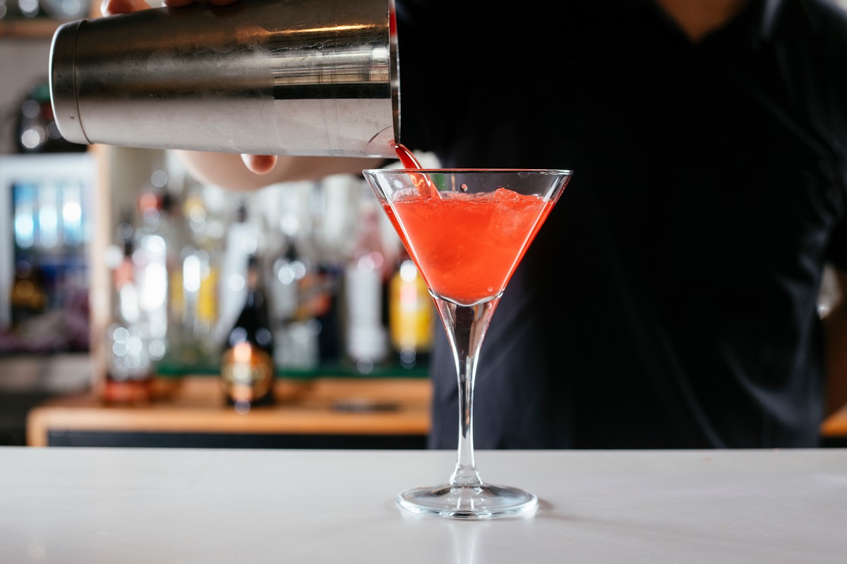 closeup cu un barman care toarna un cocktail cosmopolitan intr-un pahar asezat pe bar