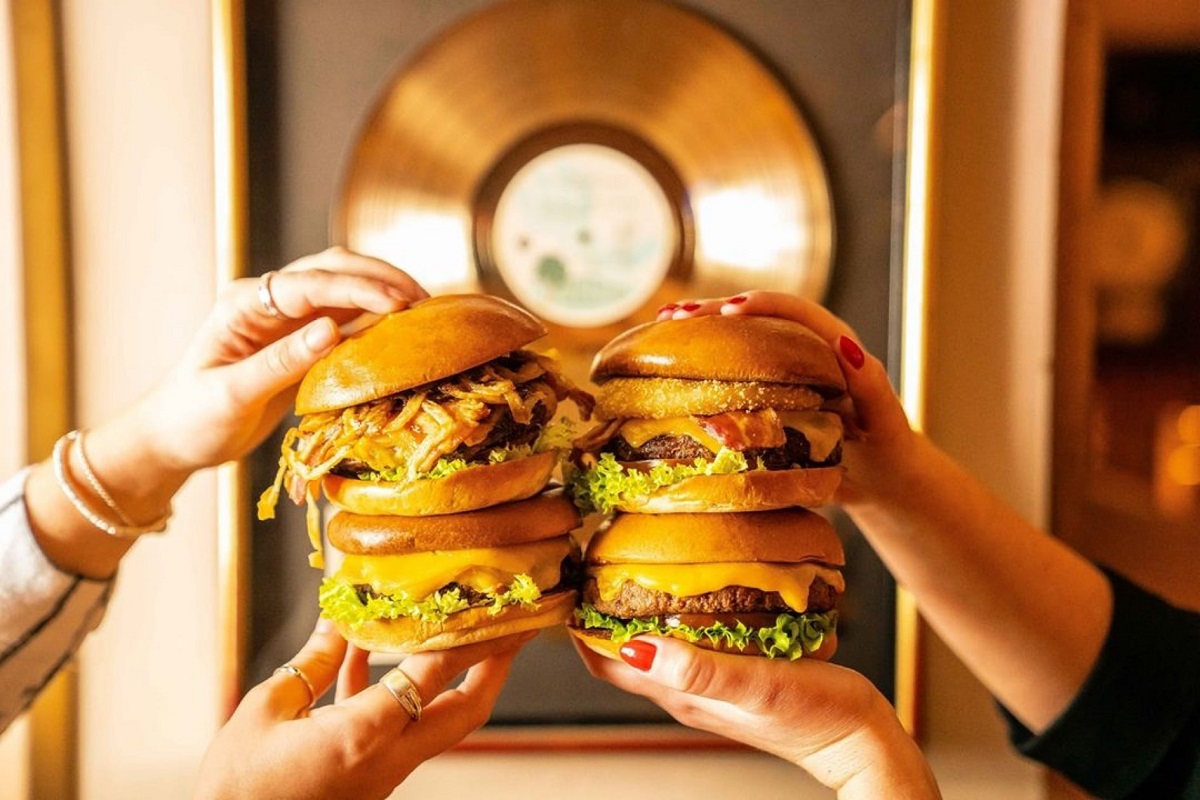 close up cu doi burgeri foarte inalti, pusi unul langa altul, tinuti in mana de doua persoane 