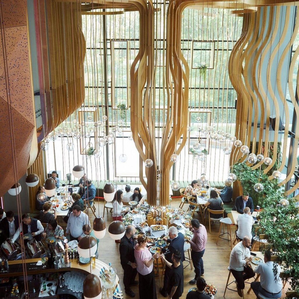 restaurant cu tavan inalt si element de design in mijloc, si clienti care iau masa de pranz la restaurante pipera