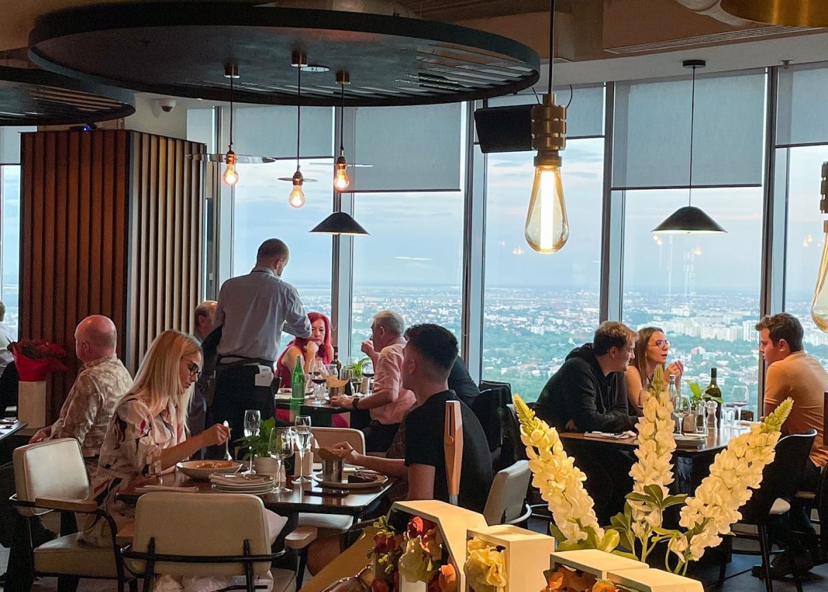 restaurantul Nor cu mese pline de clienti care iau masa, ospatar care serveste, iar in fundal ferestre mari cu panorama orasului 