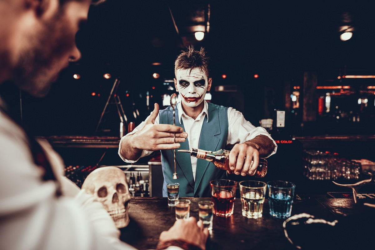 barman in costum de Halloween, pictat pe fata ca Joker din Batman, turnand bauturi in pahare clientilor la petrceri de Hallloween