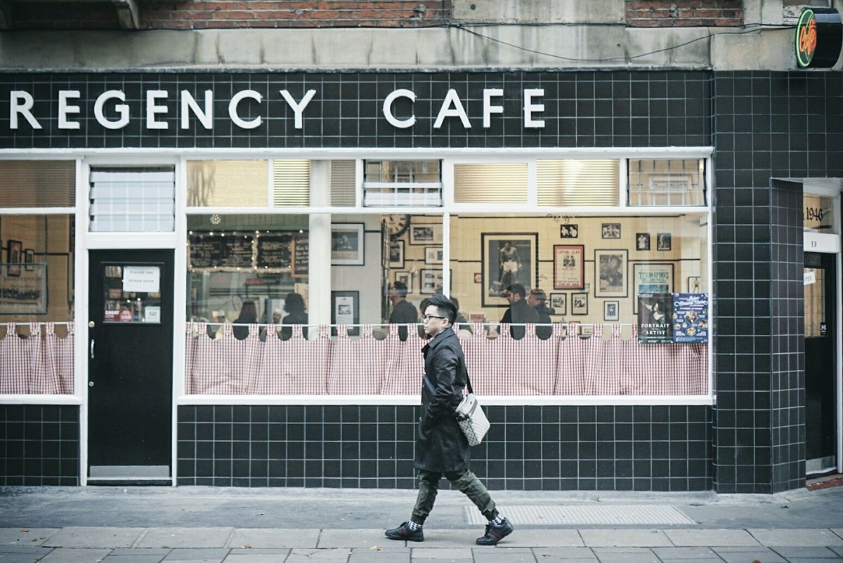 barbat asiatic care trece prin fata cafenelei Regency Cafe din Londra, cu pereti vopsiti negru, geamuri mari si perdelute la geam. Un loc unde mîncăm în Londra