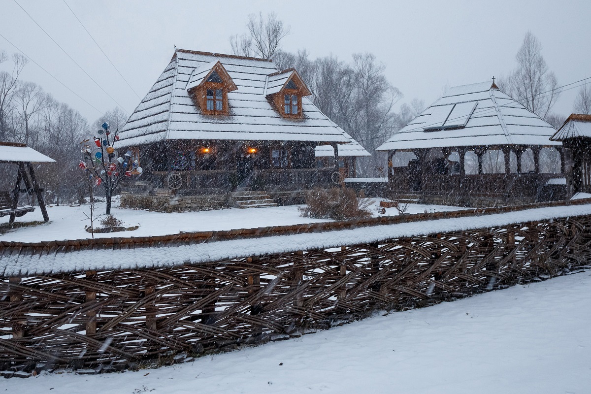 2 cabanute mici din lemn, cu gard din nuiele, aramuresean, fotografiate in timp ce ninge afara, acoperite de zapada, la Casuta din Scobai, destinație experiență de Crăciun