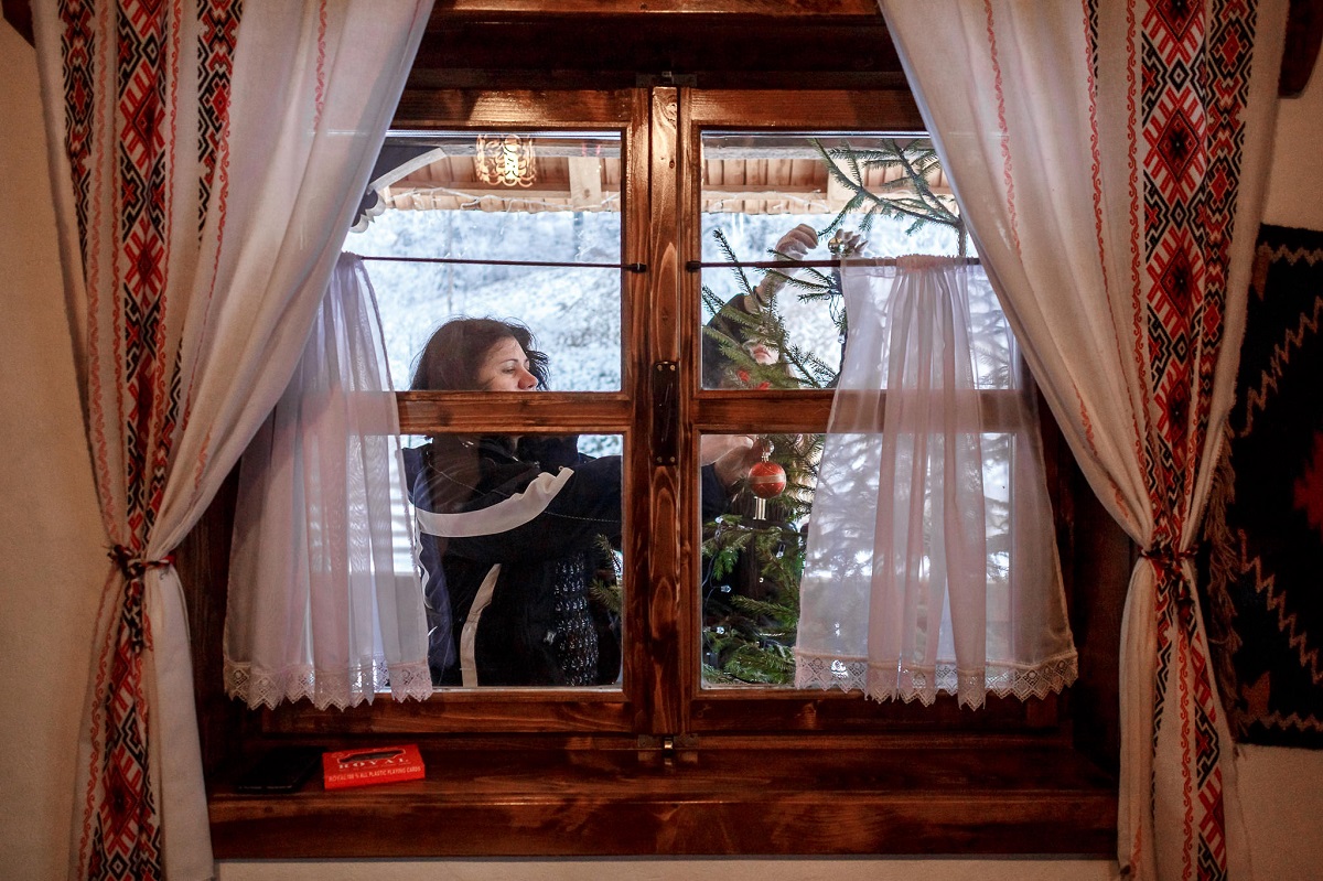 femeie fotografiata prin dreptul unei ferestre cu perdelute traditional romanesti, in  timp ce impodobeste bradul in pridvorul casei, la Casuta din Scobai, destinație experiență de Crăciun