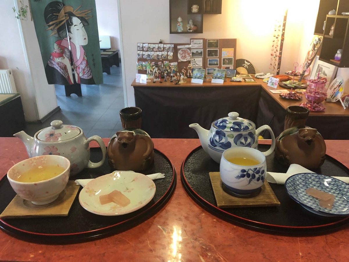 Ceainăria cu specific japonez Sakura din Cluj, fotografie de la interior, cu tavi negre pe care sunt cesti si ceainice, iar in fundal dulap cu diverse tipuri de ceai la pliculeț, si un poster atarnat, textil, pictat cu o japoneza
