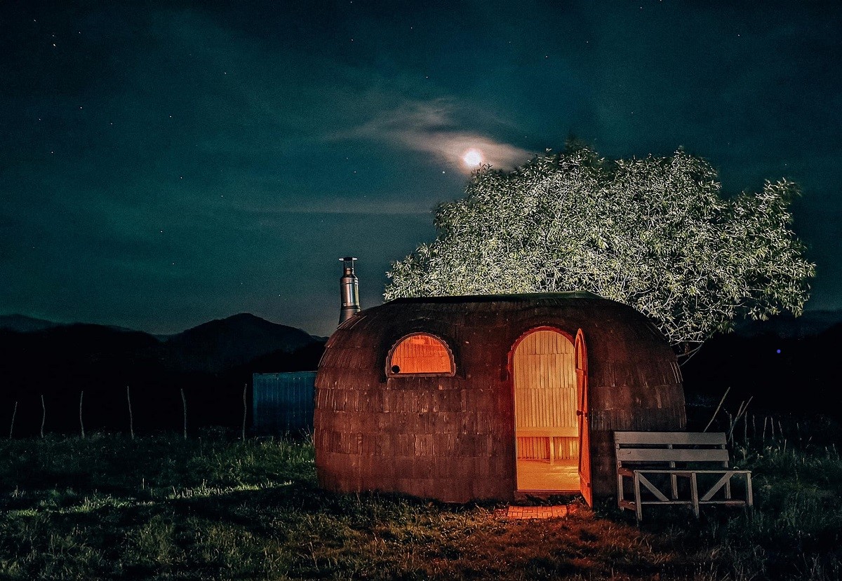 sauna de la Ursa Mica, din lemn, fotografiata noaptea, cu lumina aprinsa si un copac luminat in spate iar deasupra cerul cu luna plina 