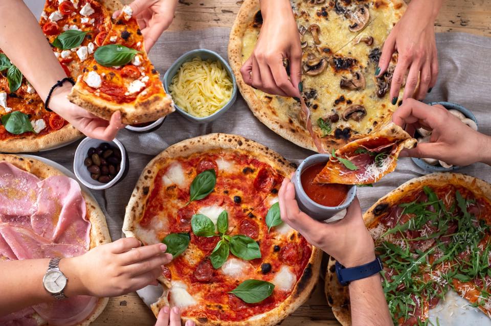 4 tipuri de pizza asezate pe masa, din care mananca mai multe persoane, cu mici boluri de sos si branza rasa, fotografiate de sus