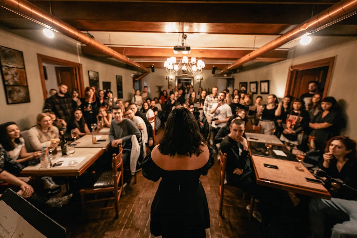 seara de poezie la Arte Cafe Iași, unde o femeie in rochie neagra e fotografiata din spate in timp ce citeste o poezie cu fata catre sala plina de oameni asezati la mese