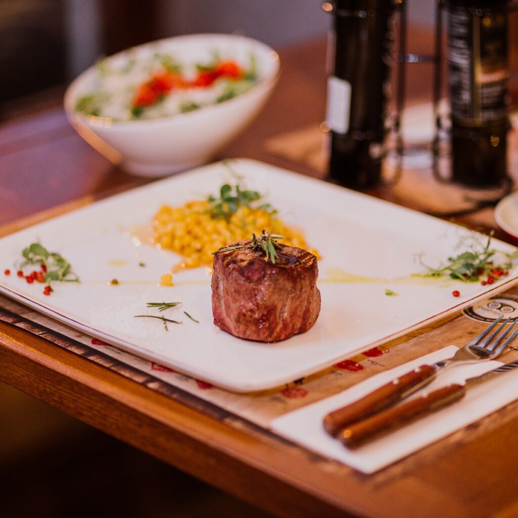 steak de vita servit pe farfurie alba, cu crenguta de rozmarin, si garnitura in fundal