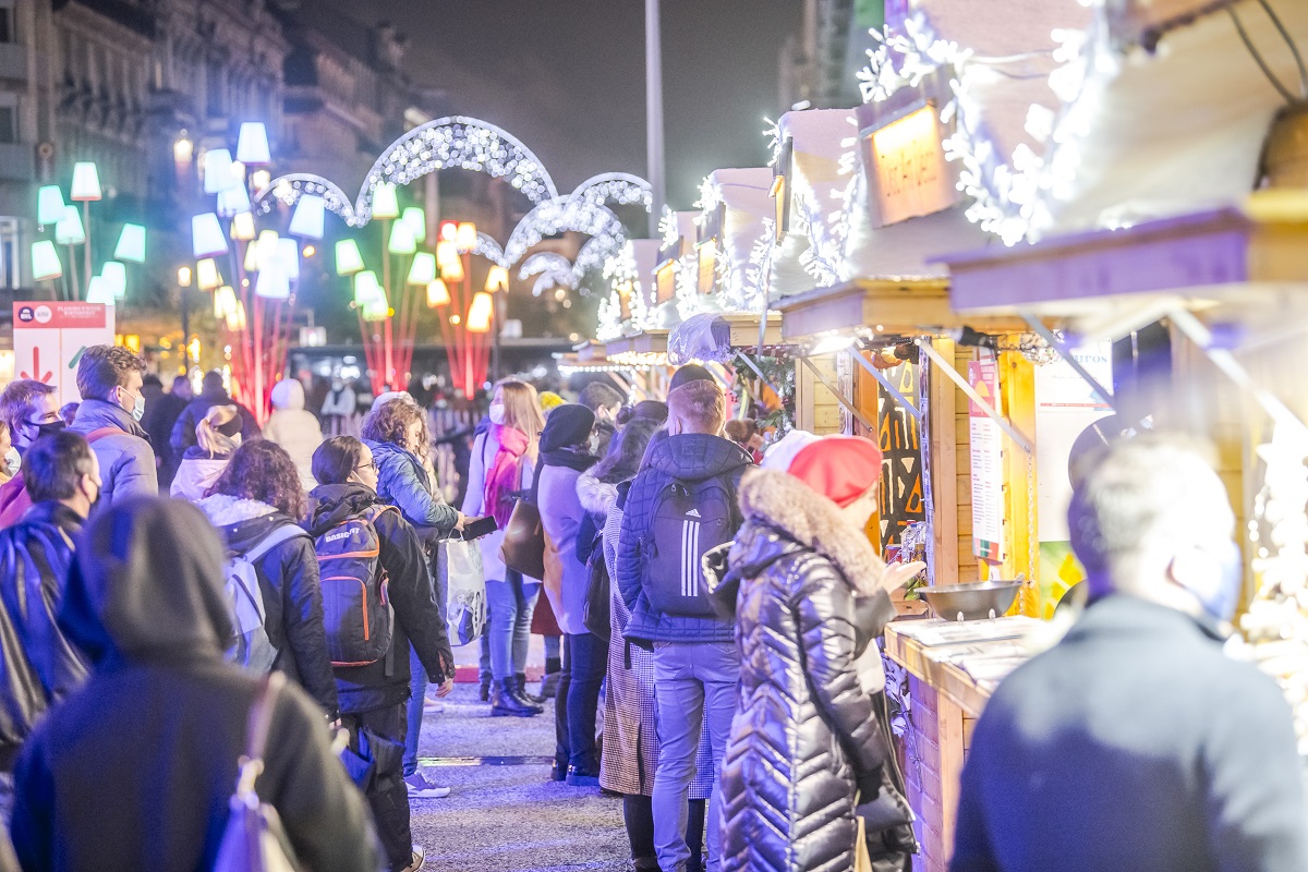 Piata de Crăciun de la Bruxelles, fotografiata seara, aglomerata cu oameni care fac cumparaturi la casutele de lemn, iar in jur multe decorațiuni de Crăciun